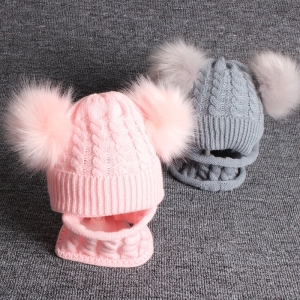 twee pompomhoeden met geïntegreerde sjaal, één roze aan de voorkant en de andere grijs aan de achterkant rechts, op grijze stof