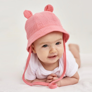 Liggend babymeisje op een witte honingraatstof met een wit rompertje en een roze mousseline mutsje met oren en koord.